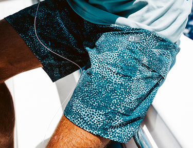 Close-up of a person sitting outdoors, wearing teal patterned shorts and a light-colored shirt. The shorts have a small label and a speckled design, and sunlight highlights the texture of the fabric.