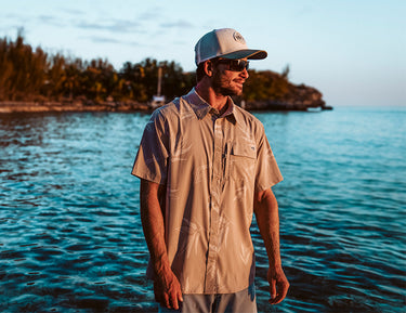 A man wearing sunglasses, a beige short-sleeve shirt, and a baseball cap stands in shallow ocean water near a forested shoreline at sunset.