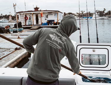 A person in a green Salty Crew hoodie stands on a boat, holding a pole near a fishing harbor, with a fish market graphic on their back and fishing rods visible in the background.