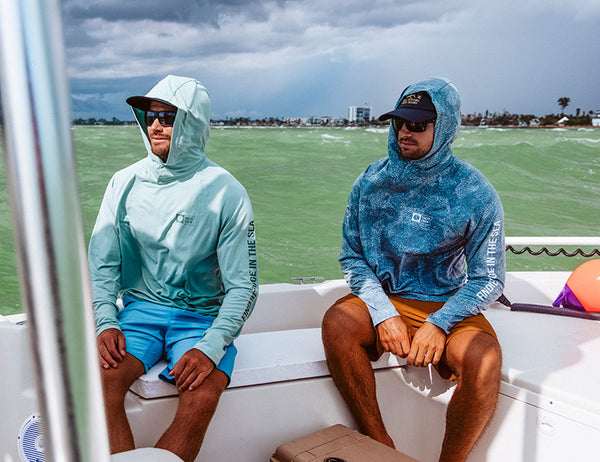 Two men wearing hooded sun shirts, sunglasses, and shorts sit on a white boat with light green water and a cloudy sky in the background. One shirt is light blue and the other is dark blue.