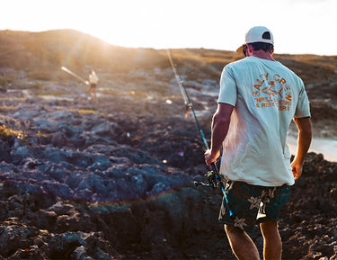 A man wearing a white Salty Crew t-shirt, floral shorts, and a cap walks over rocky terrain holding a fishing rod. Another person with fishing gear is seen in the background at sunset.