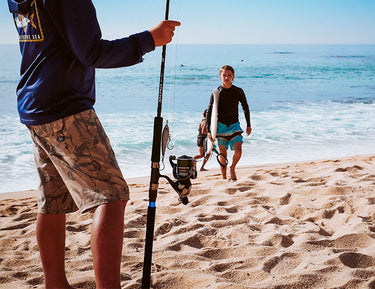 Two boys walk up a sandy beach toward another person holding a fishing rod. The ocean waves are in the background under a clear blue sky.