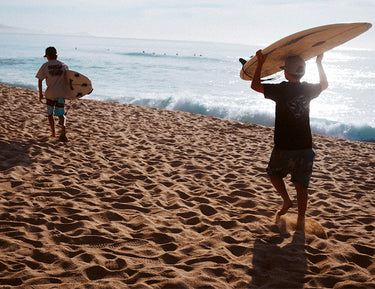 Two people walk on a sandy beach toward the ocean, each carrying a surfboard under one arm. The sun is bright, and small waves are visible in the background.