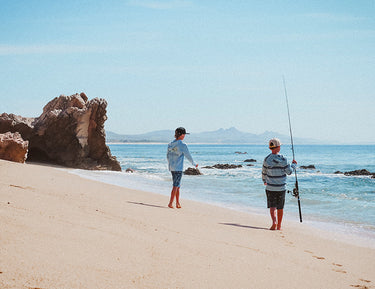 Two boys walk along a sandy beach near rocks; one carries a fishing rod. The ocean is calm, and there are distant mountains under a clear blue sky.