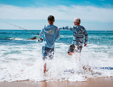 Two boys stand at the shoreline with ocean waves splashing around them; one holds a fishing pole while both look toward the water under a bright, blue sky.