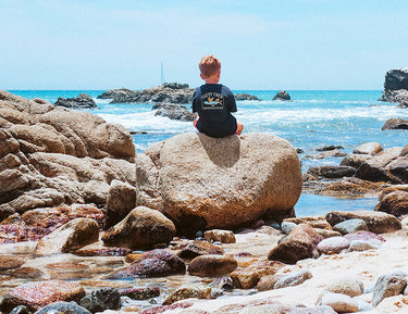 A young boy with red hair sits alone on a large rock by the ocean, facing the waves. Rocky tide pools and blue sky surround him, creating a serene beach scene.
