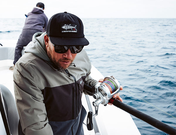 A man wearing sunglasses, a cap, and a gray jacket is fishing on a boat, holding a fishing reel over the side. Another person faces away, standing at the back of the boat. The ocean surrounds them.