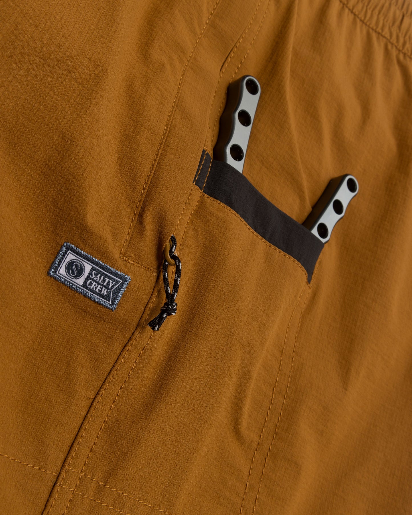 Close-up of Salty Crew Crew Boatshort Ewaist in Bronze Brown, made with quick-dry ripstop fabric, featuring a zippered pocket with branded patch and a black-edged pocket holding two gray-handled multitools.