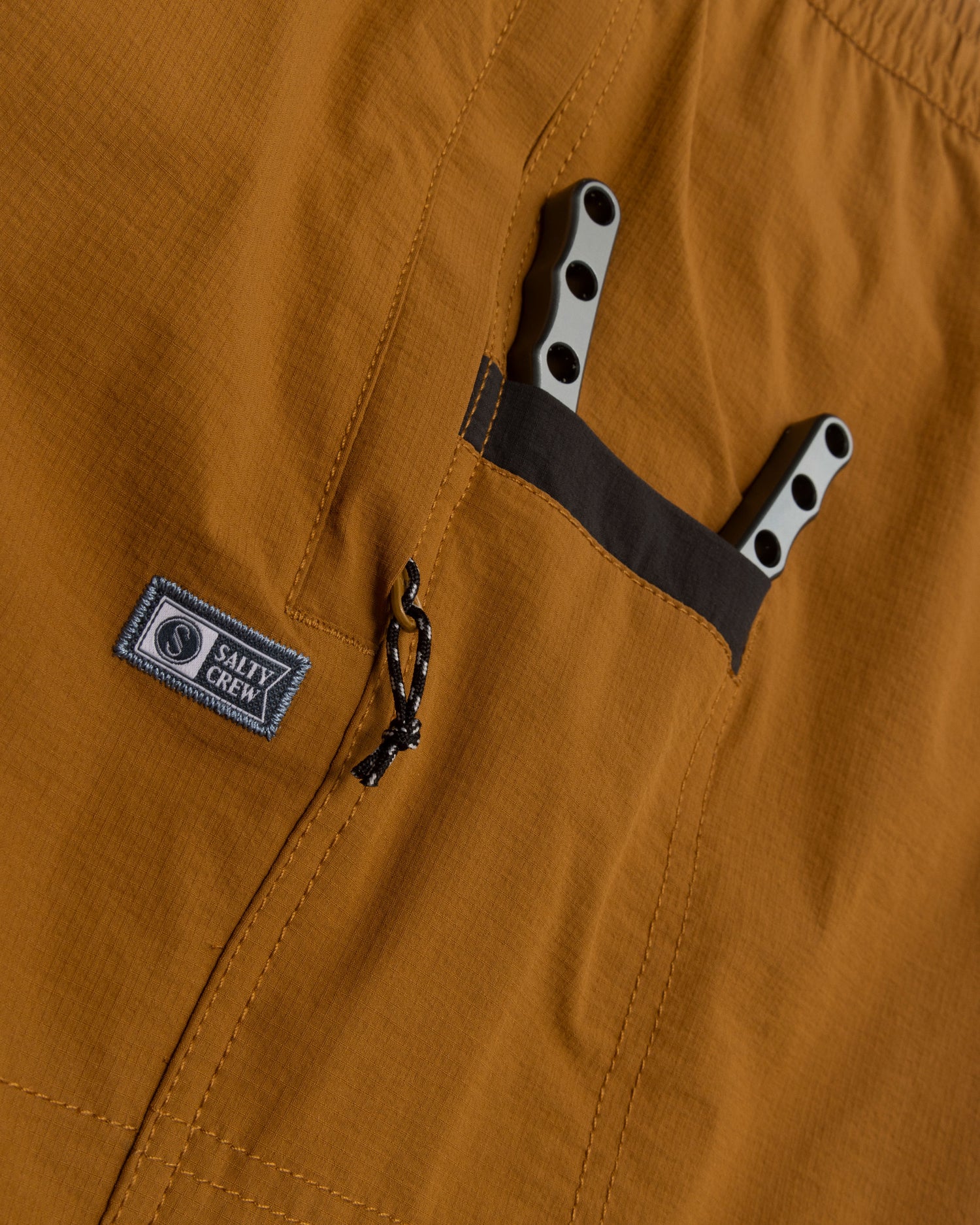 Close-up of Salty Crew Crew Boatshort Ewaist in Bronze Brown, made with quick-dry ripstop fabric, featuring a zippered pocket with branded patch and a black-edged pocket holding two gray-handled multitools.