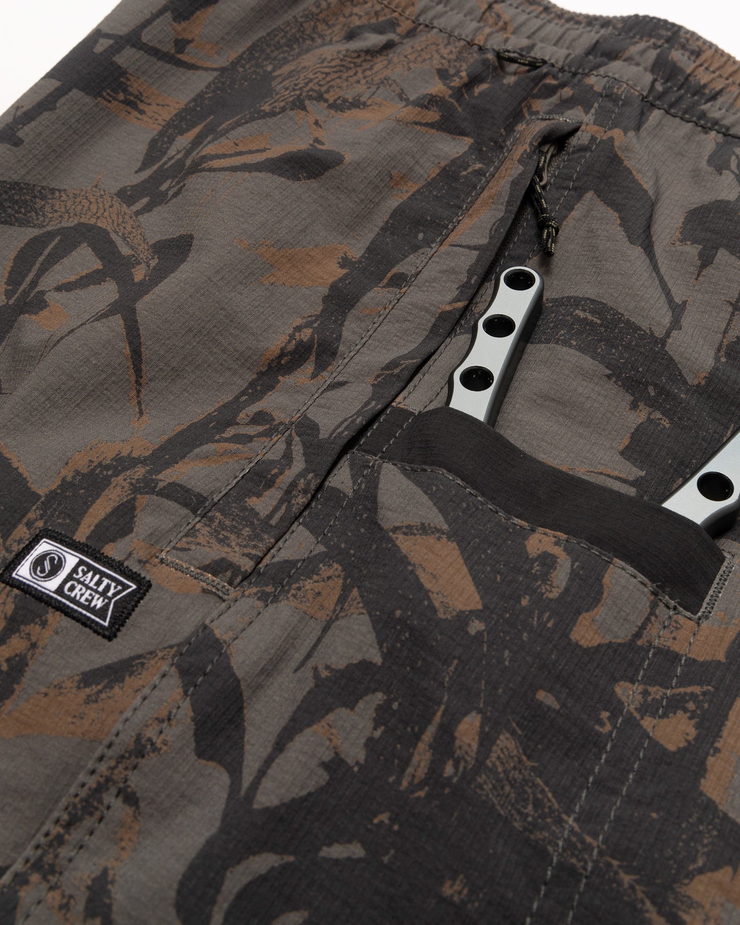 Close-up of Salty Crew Crew 18" Ewaist Boatshort in Dark Kelp Camo, featuring durable ripstop fabric and a label near the pocket. A silver butterfly knife is partially visible.