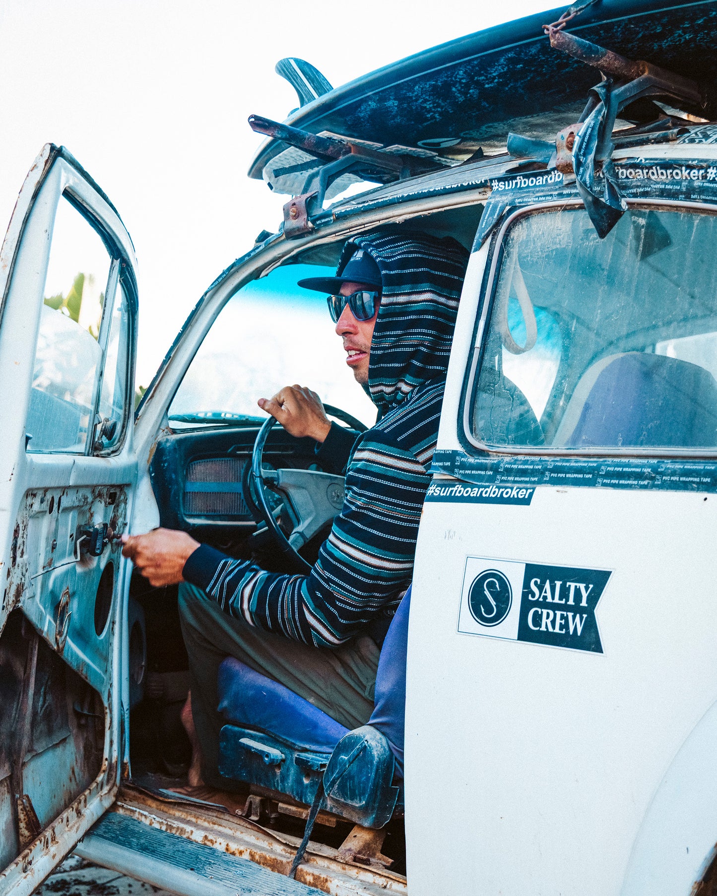 Wearing the Salty Crew Crewser Fleece Hood in Pirate Black and sunglasses, a person sits in an old white car with surfboards on the roof, opening the door that features a Salty Crew sticker.