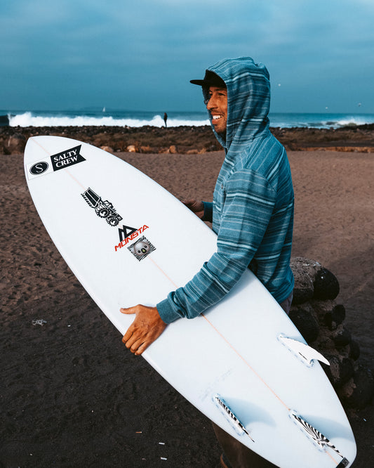 A man wearing the Salty Crew Crewser Fleece Hood in Mid Blue holds a white surfboard covered in stickers, standing on a sandy beach with waves and rocks in the background beneath a cloudy sky.