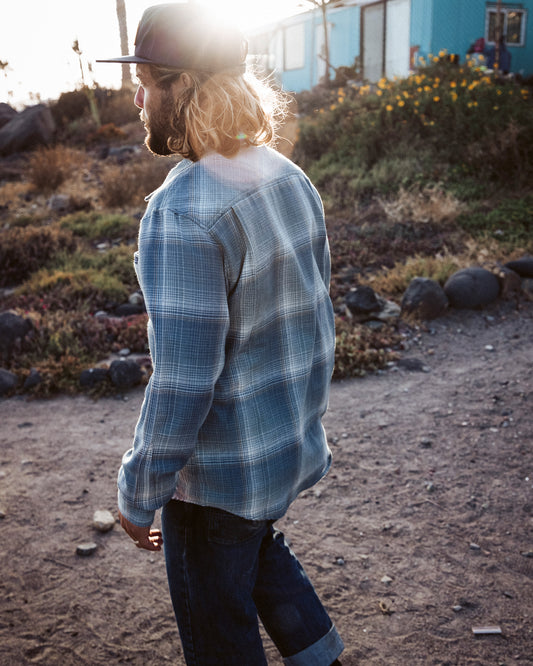 A person with long hair and a cap walks outdoors on a dirt path, wearing the Salty Crew Early Bird Flannel - Trooper and jeans. Sunlight shines from behind, creating a soft, warm glow as rocks and plants line the path. The lightweight twill fabric of the Early Bird Flannel offers comfort for an easy stroll.