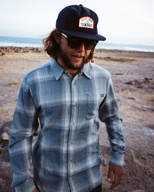 A man with long hair, wearing sunglasses, a dark cap, and the Salty Crew Early Bird Flannel - Trooper—a yarn-dyed plaid shirt with a soft hand garment wash—stands outdoors on rocky ground near the ocean, looking slightly downward with a relaxed expression.