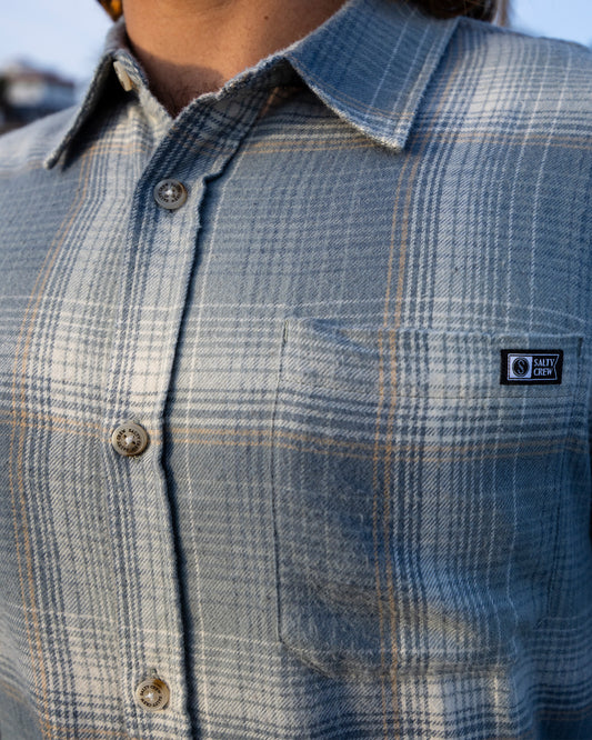 A person is seen wearing the Salty Crew Early Bird Flannel in Trooper, a blue and gray yarn-dyed plaid button-up shirt featuring a logo patch on the chest pocket. Made from lightweight twill with a soft hand garment wash, the shirt is buttoned up and only the torso is visible.