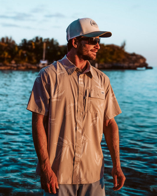 A man in a Salty Crew Flagship UV Button Up - Dark Khaki, sunglasses, and a white cap stands by the water at sunset with trees and a boat behind him. The shirt features UPF 50+ sun protection and moisture-wicking fabric.