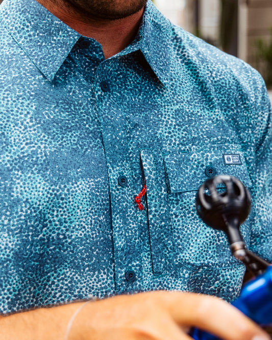 A person wearing the Salty Crew Flagship UV Button Up in Dark Navy (UPF 50+) holds equipment with a black circular part in the foreground, showing only their lower face, chest, and hand.