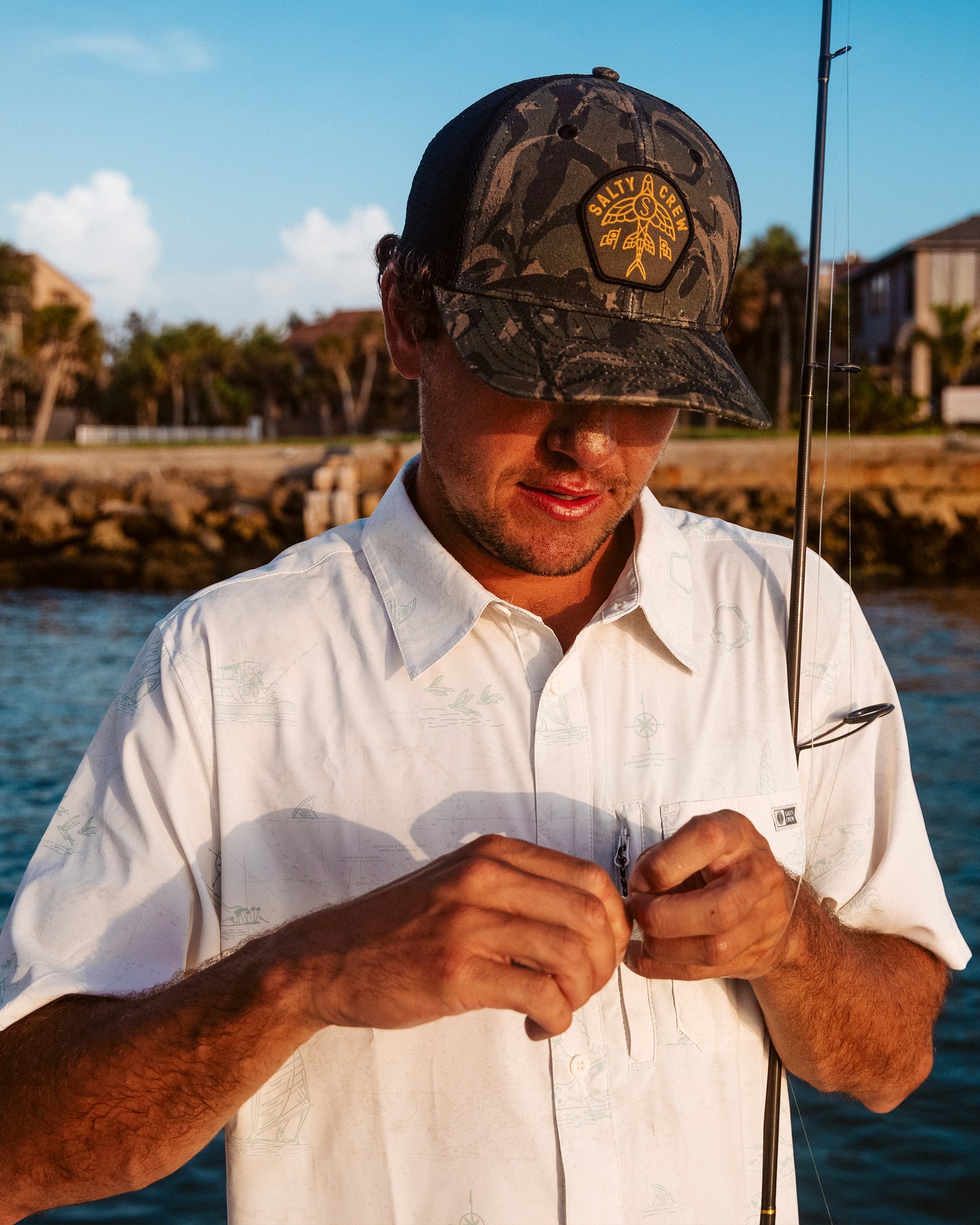 A man in a Salty Crew Flagship UV Button Up - White stands by the water, focused on tying a fishing hook. Houses, rocks, and palm trees add to the scenic backdrop.