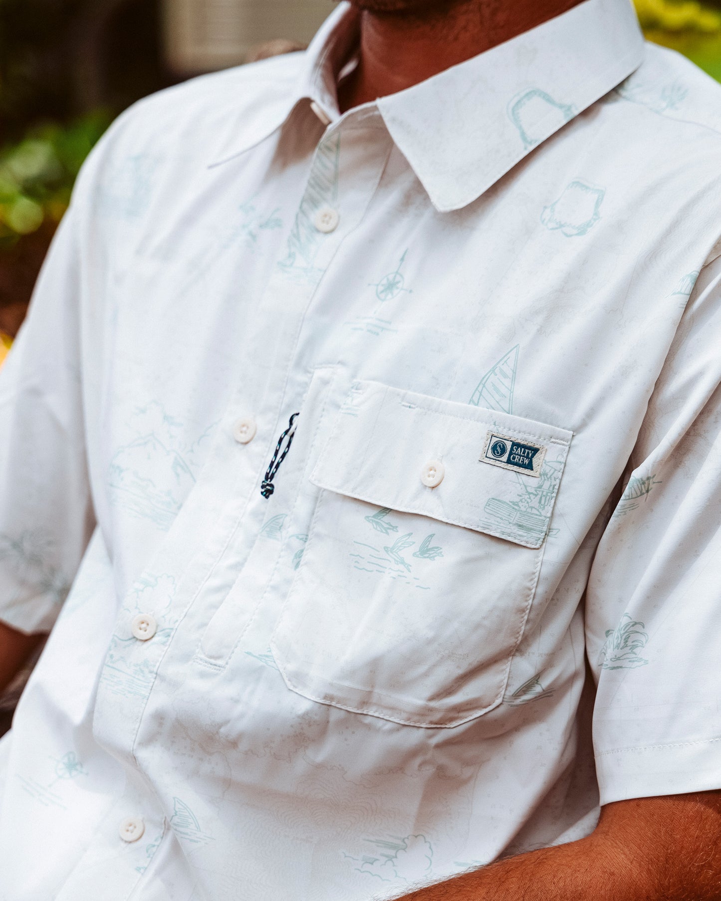 A person wears the Salty Crew Flagship UV Button Up in white, featuring a subtle light blue nautical print, chest pocket with logo patch, and quick-dry fabric for comfort on warm days. Face is not visible.