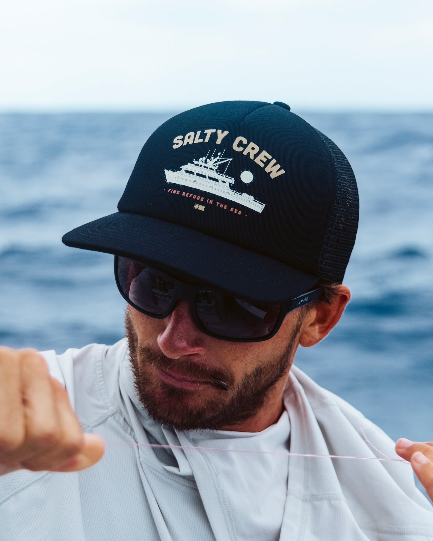 A man in sunglasses and a Salty Crew Good Times Foamie Trucker - True Black, featuring a fishing boat graphic, is outdoors holding a fishing line with the ocean blurred in the background.