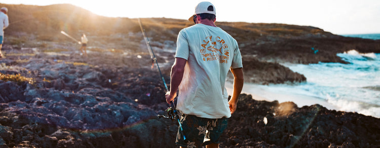 A person wearing a white cap and T-shirt carries a fishing rod while walking on rocky terrain by the ocean, with the sun shining in the background.