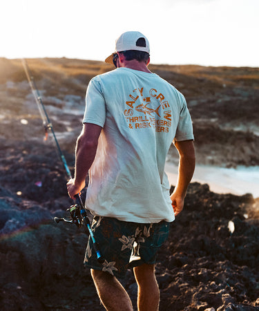A person wearing a white Salty Crew t-shirt, patterned shorts, and a cap holds a fishing rod while walking on rocky terrain near the water at sunset.