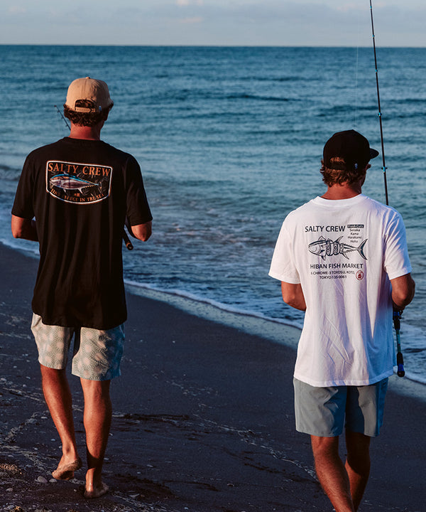 Two people wearing shorts, T-shirts, and hats walk barefoot along a sandy beach beside the ocean; one is holding a fishing rod. The T-shirts have Salty Crew logos on the back.