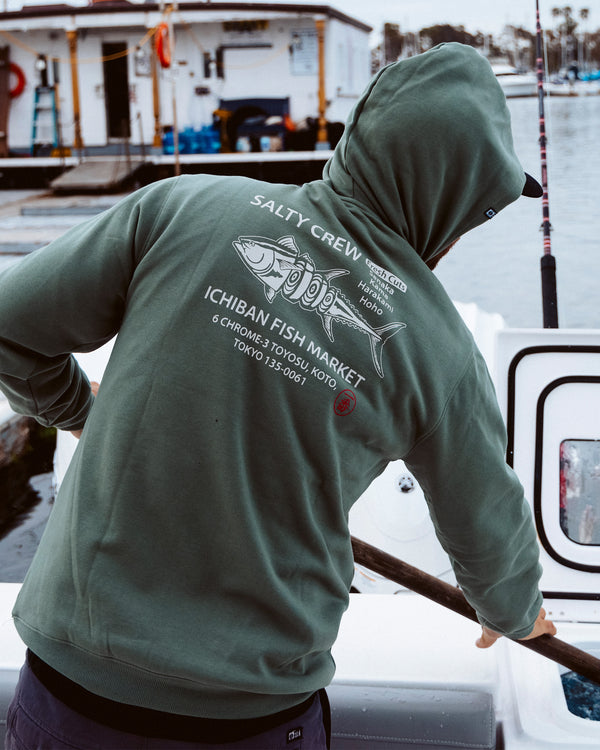 A person wearing a green Salty Crew hoodie with a fish graphic and Ichiban Fish Market text stands on a boat docked by the water, holding a fishing rod.