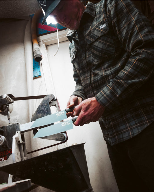 A person in a plaid shirt and baseball cap sharpens a limited edition Salty Crew Leucadia x Salty Crew 8” Hamachi Fillet Knife with Detroit Diesel Green cerakote on a bench grinder in a tool-filled workshop.