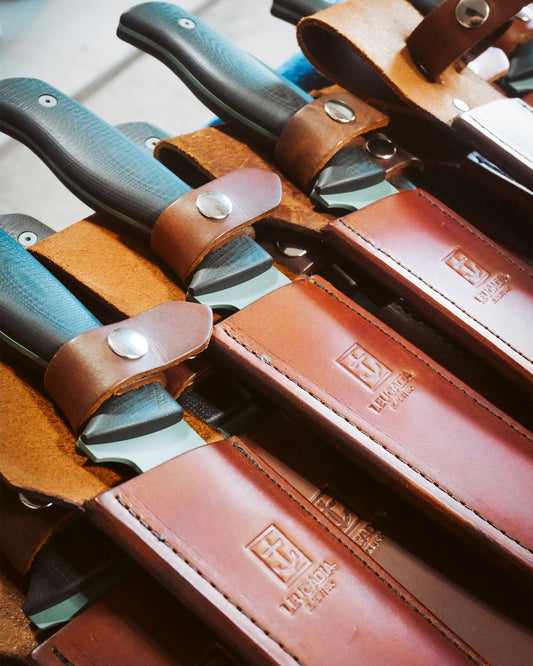 Several Salty Crew knives with dark handles, including a limited edition Leucadia x Salty Crew 8” Hamachi Fillet Knife, are secured in brown leather sheaths with stamped logos and metal snaps, neatly arranged in rows.