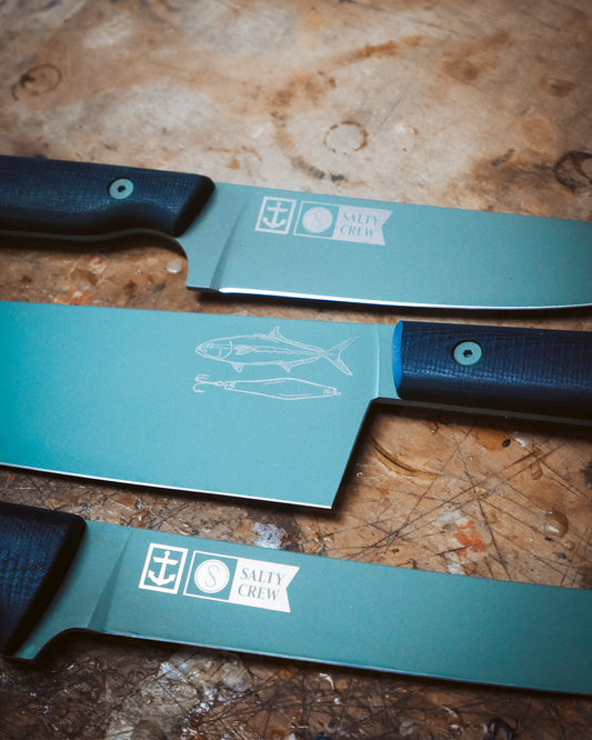 Three Leucadia x Salty Crew 5” Snapper Utility Knives with dark handles lie on a scratched surface. Their Detroit Diesel Green blades, engraved with a fish, anchor, and Salty Crew logo, create a striking limited edition set.