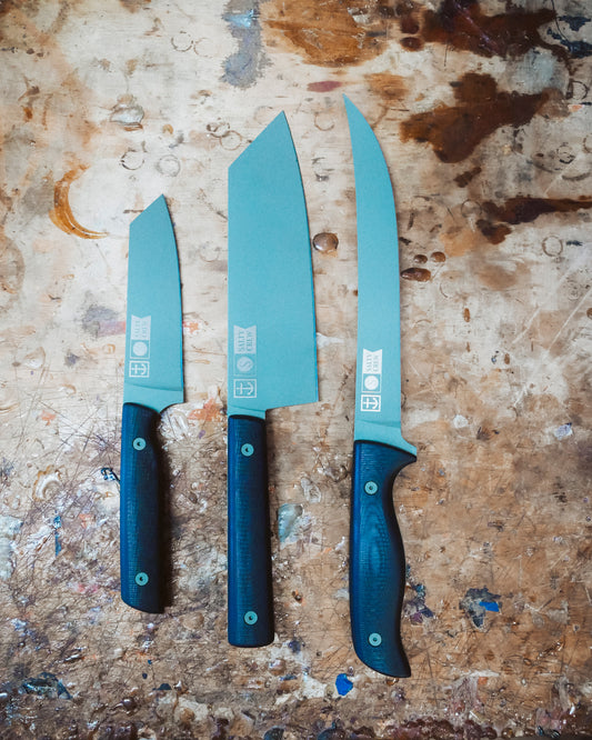 Three Salty Crew kitchen knives with black handles and silver blades, including the Leucadia x Salty Crew 5” Snapper Utility Knife (limited edition), are arranged side by side on a stained, textured wooden surface. The knives differ in blade shape and size.