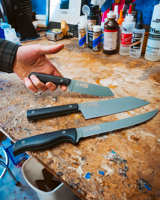 A person holds a Salty Crew Leucadia x Salty Crew 5” Snapper Utility Knife with a black handle above a wooden workbench, where two more knives rest amid scattered bottles, tools, and supplies in the background.