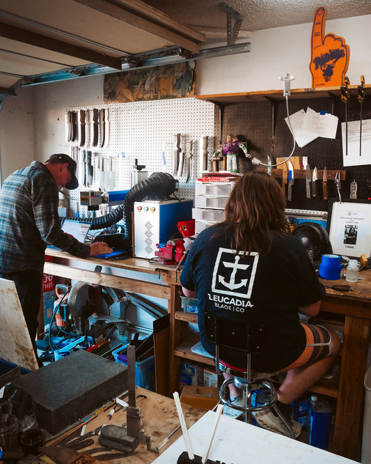 Two people work at a busy workshop bench with tools. One wears a Leucadia Blade Co. shirt, while the other stands, focused on the Salty Crew Leucadia x Salty Crew 5” Snapper Utility Knife next to a handmade leather sheath.