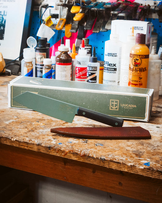 The Leucadia x Salty Crew 7” Bunka Knife by Salty Crew, featuring a green handle and AEB-L Swedish stainless steel blade, rests on its box amid adhesives and tools, with a brown leather sheath displayed in front.