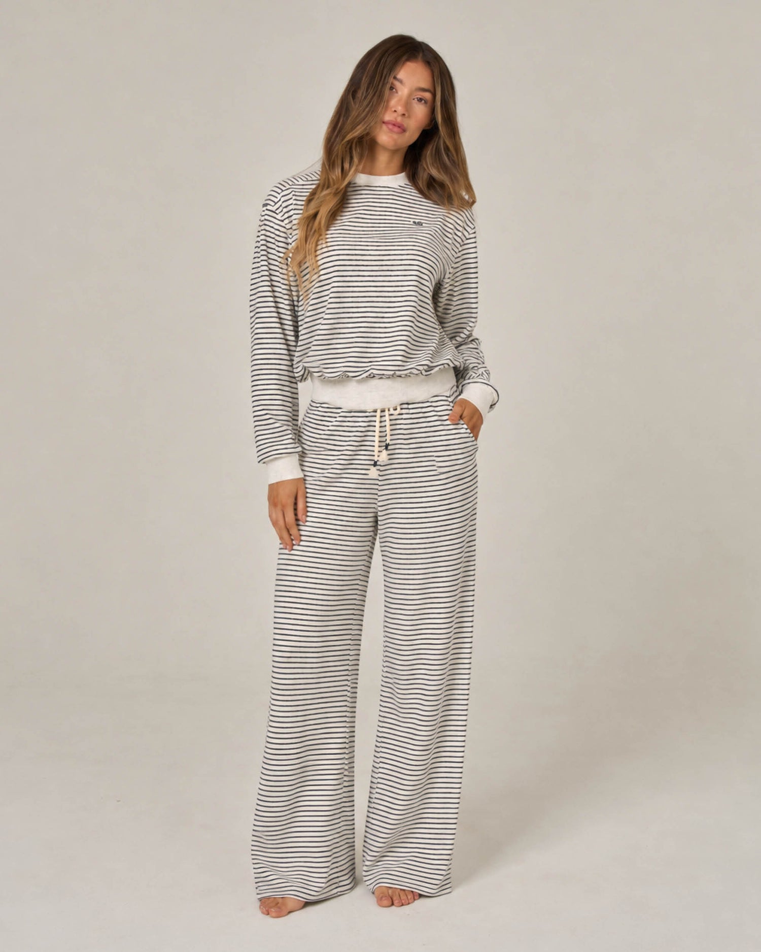 A person stands barefoot, wearing Salty Crew’s Leeward Pant in Light Heather paired with a matching striped long-sleeve top. The outfit, made from a soft cotton-polyester blend, is loose-fitting and casual, featuring a crew neck Terry Stripe pullover with drawstrings at the waist. The background is plain and neutral.