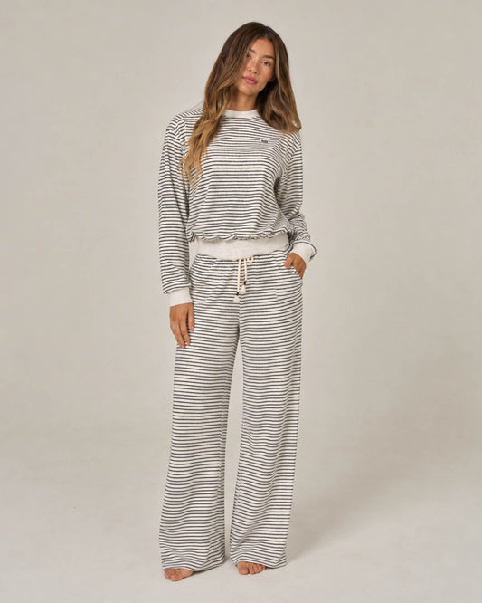 A person stands barefoot, wearing Salty Crew’s Leeward Pant in Light Heather paired with a matching striped long-sleeve top. The outfit, made from a soft cotton-polyester blend, is loose-fitting and casual, featuring a crew neck Terry Stripe pullover with drawstrings at the waist. The background is plain and neutral.