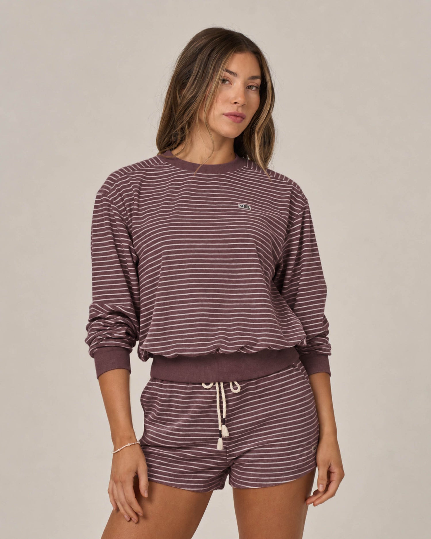 A woman stands against a plain light background wearing the Salty Crew Leeward Crew in Maroon—a matching set featuring a crew neck long-sleeve top and drawstring waist shorts, crafted from soft cotton-polyester pullover fabric with mauve and white stripes.