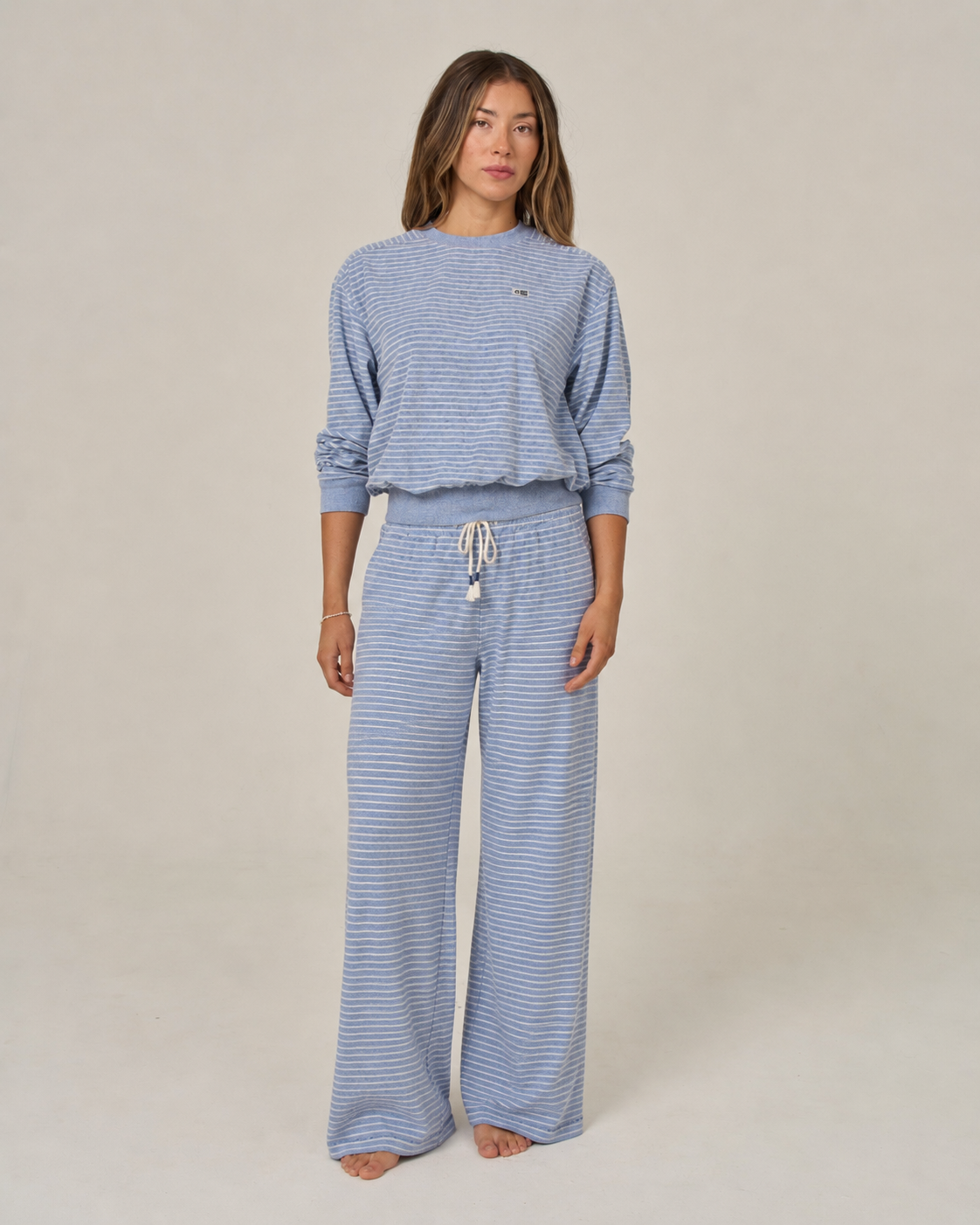 A woman stands barefoot against a plain background, wearing the Salty Crew Leeward Pant in Blue Heather—a pair of wide-leg lounge pants with a drawstring waist, crafted from a soft cotton polyester blend.