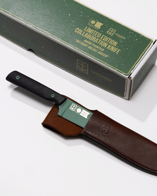 The Salty Crew Leucadia x Salty Crew 5” Snapper Utility Knife features a black handle, Detroit Diesel Green blade, and comes partially sheathed in handmade leather, shown beside its green and white box on a white background.