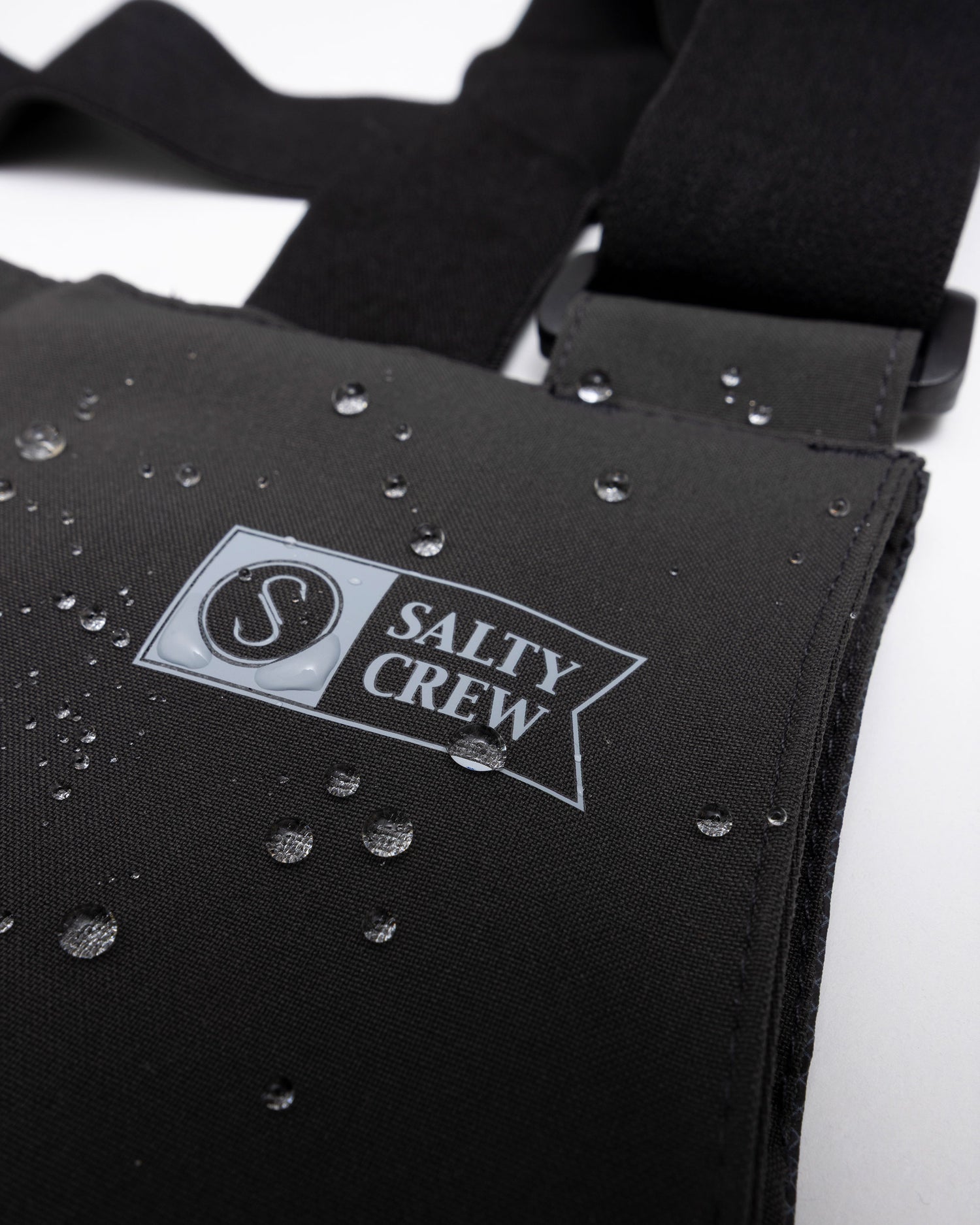 Close-up of the Long Range Tech Bib - Black by Salty Crew, showing its ultra-waterproof black fabric strap with water droplets and a Salty Crew logo—a top choice for fishing gear.
