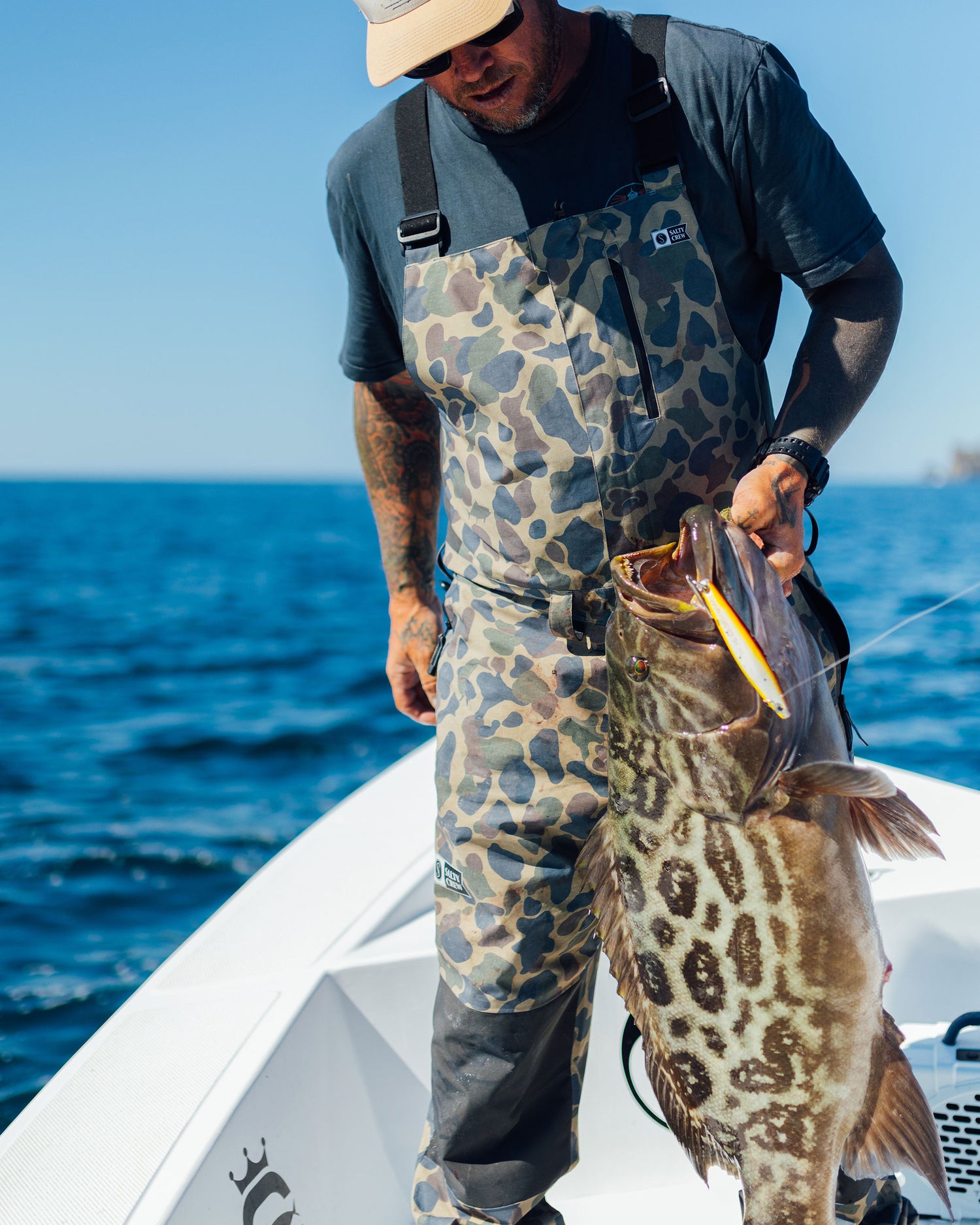 Wearing Salty Crew's Long Range Tech Bib in Sand Camo, a person stands on a boat holding a large spotted fish, with the ocean and clear blue sky in the background.