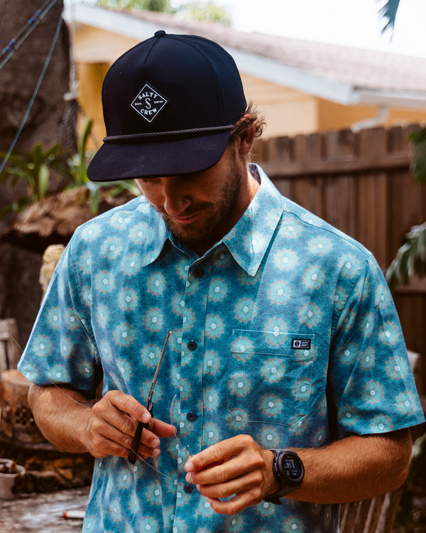 A man wearing the Salty Crew Crew Button Up - Slate and a dark baseball cap stands outdoors near a wooden fence, holding thin tools with greenery and a house in the background.