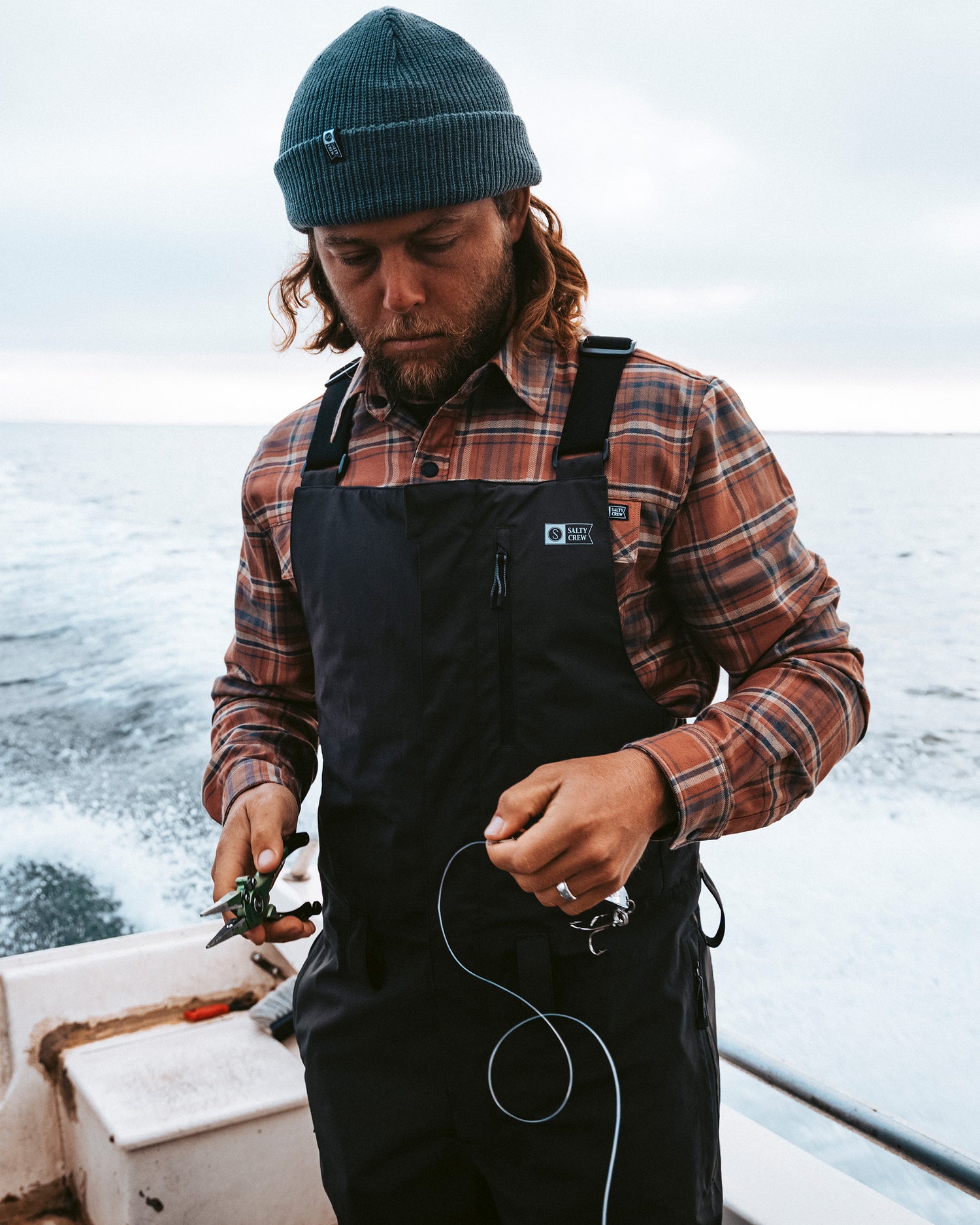 Wearing the Salty Crew Long Range Tech Bib in black, a man stands on a boat in front of the ocean and cloudy sky, working with fishing line and pliers while dressed in a blue beanie and plaid shirt.