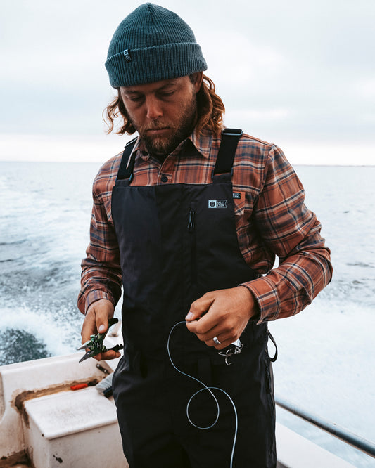 Wearing the Salty Crew Long Range Tech Bib in black, a man stands on a boat in front of the ocean and cloudy sky, working with fishing line and pliers while dressed in a blue beanie and plaid shirt.