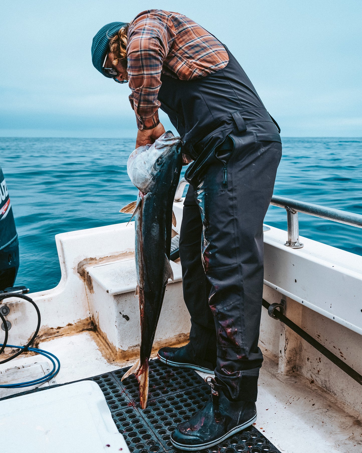 Wearing the Salty Crew Long Range Tech Bib in black, a person stands on a boat holding a large fish by the gills, with the ocean and cloudy sky in the background.