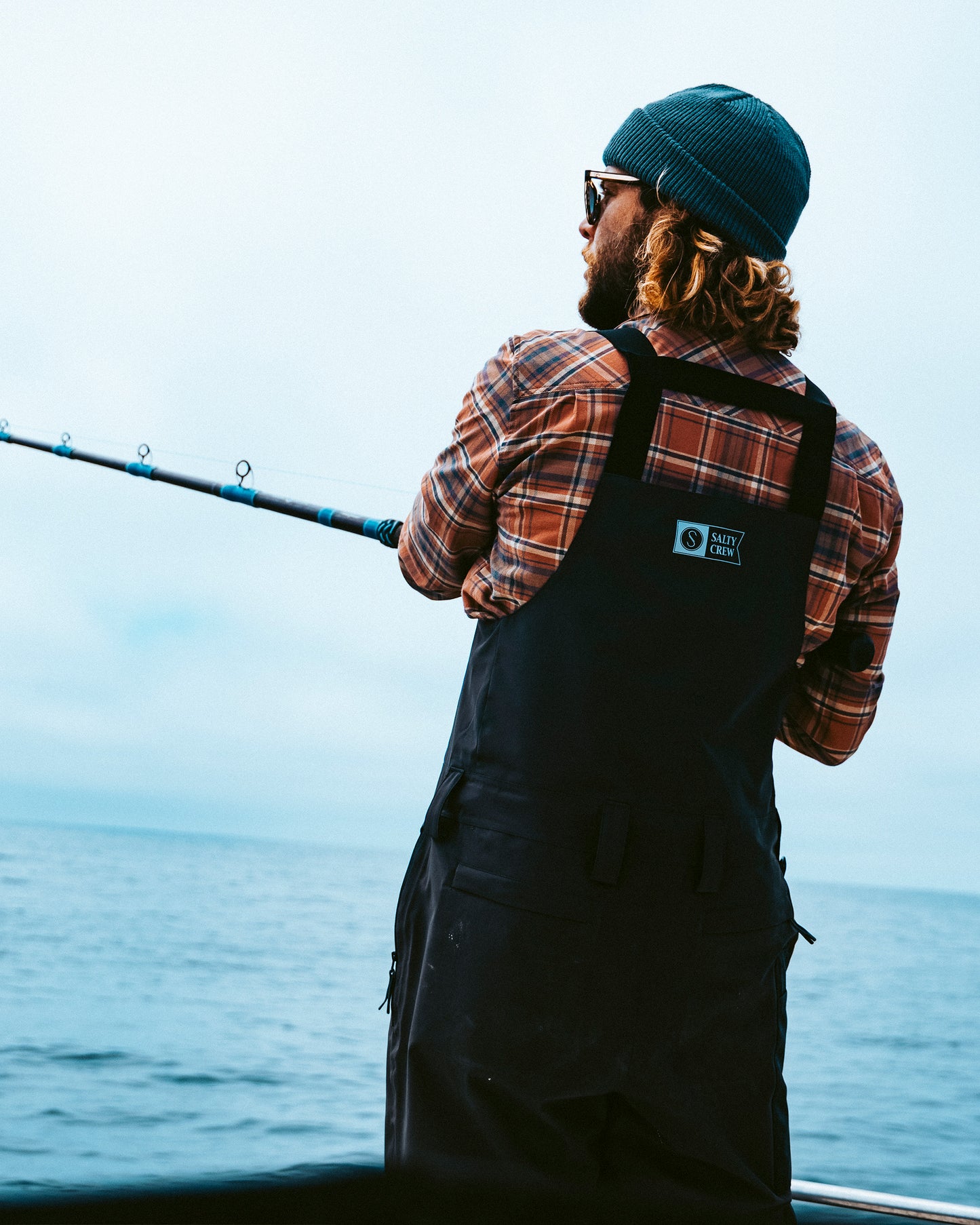 A person wearing a beanie, plaid shirt, and overalls is fishing from a boat on a calm, open body of water under a cloudy sky, outfitted in the Salty Crew Long Range Tech Bib - Black for extra protection against the elements.