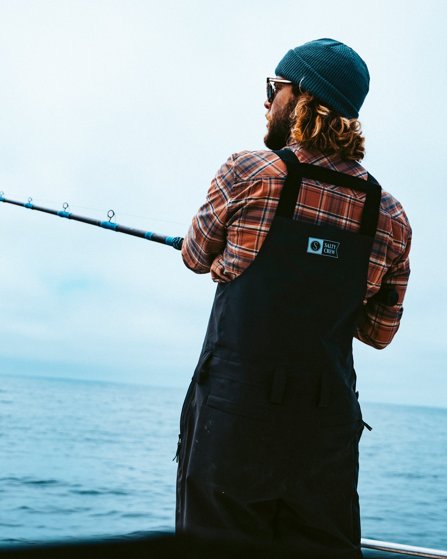 A person wearing a beanie, plaid shirt, and overalls is fishing from a boat on a calm, open body of water under a cloudy sky, outfitted in the Salty Crew Long Range Tech Bib - Black for extra protection against the elements.