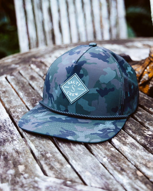 The Salty Crew Long Range Tech Snapback - Camo, featuring laser-cut breathable panels and a front logo, sits on a weathered wooden surface outdoors.