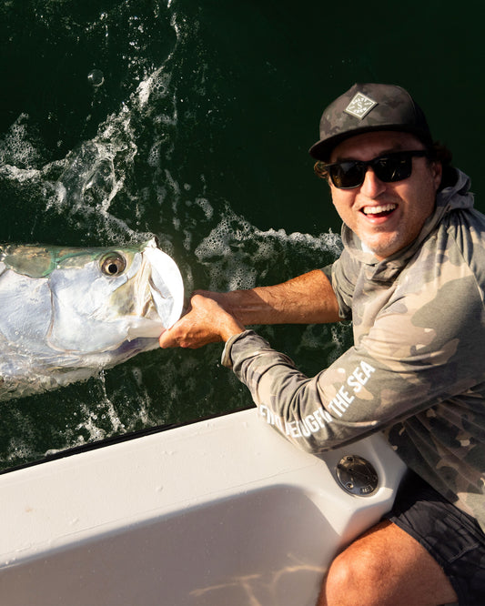A man in sunglasses, a cap, and a Salty Crew Mariner Lite UV Hoodie - Green Camo with UPF 50+ grins as he holds a large fish by the mouth over a boat side above green water.
